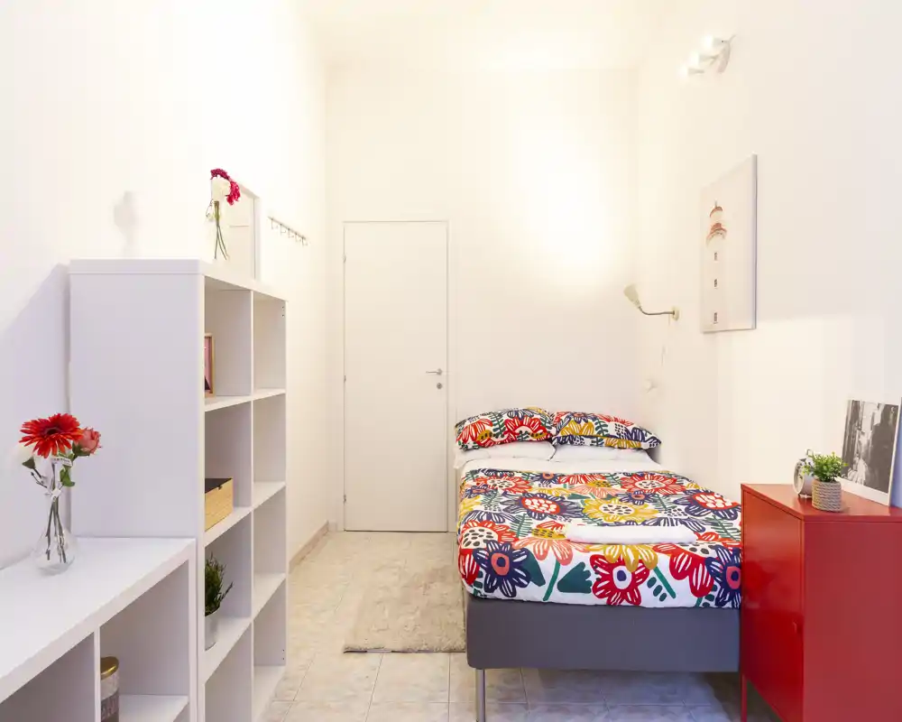 Camera da letto singola luminosa e ordinata con una coperta floreale colorata, scaffalature aperte e tocchi decorativi minimal per un’atmosfera fresca e accogliente.