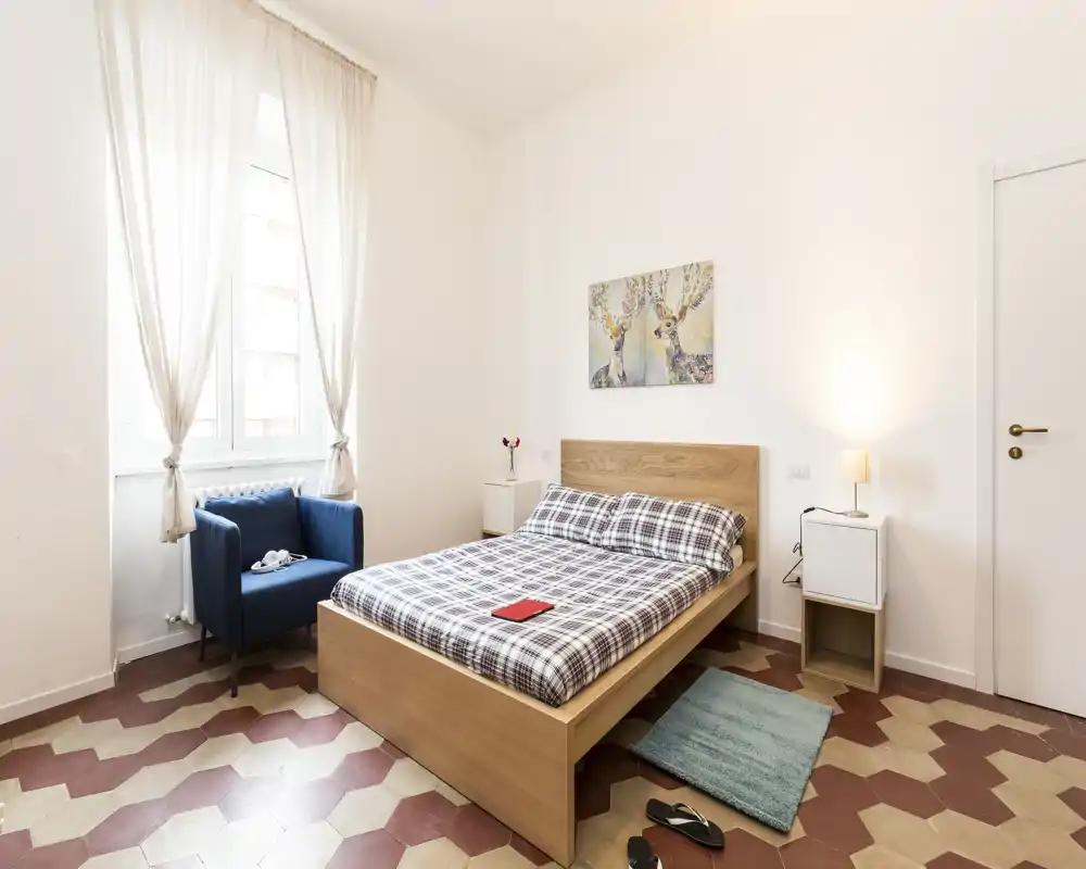 Bright, well-styled bedroom with a wooden double bed, patterned bed linen, bedside tables and a comfy blue armchair by a large window letting in abundant natural light.