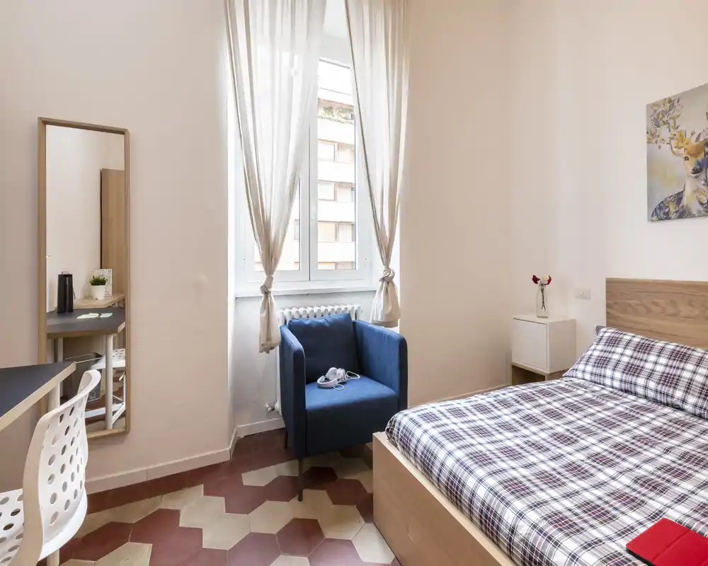 Bright, neatly styled bedroom with a double bed, cozy textiles and a blue armchair beside a large window letting in natural light.