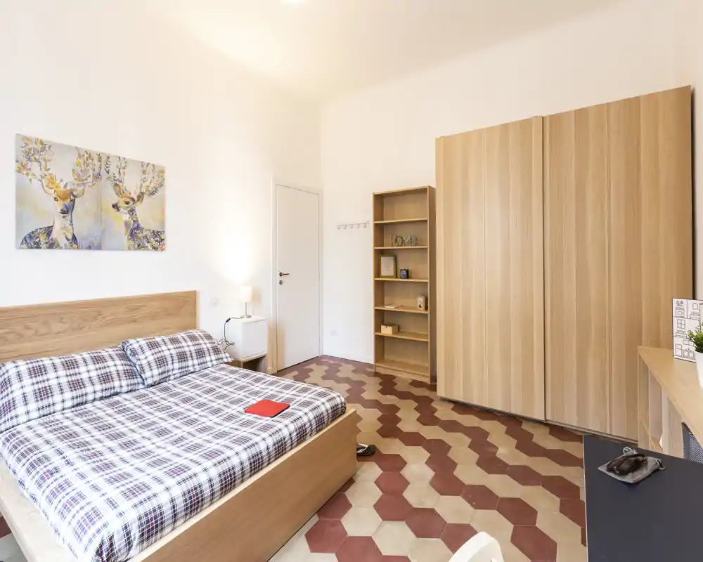Spacious, bright bedroom with a large wooden bed, patterned tile floor and built-in wardrobe. Warm decor, bedside lamp and wall artwork create a welcoming atmosphere for short or long stays.