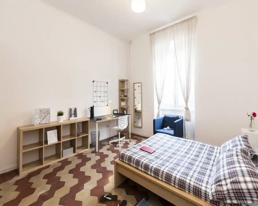 Bright, neatly styled bedroom with a double bed in the foreground, patterned floor tiles, a desk workspace and a cozy armchair by the window.
