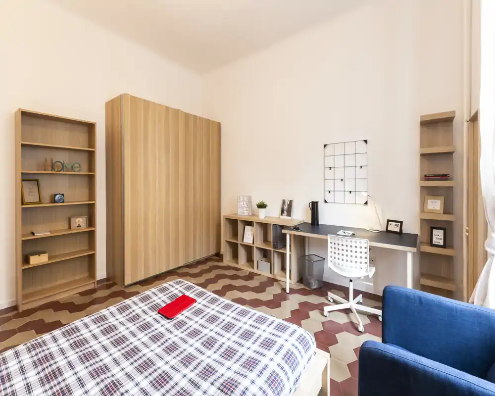 Spacious, bright bedroom with a patterned tile floor, large wardrobe, desk area and a comfortable double bed dressed in checked linens.