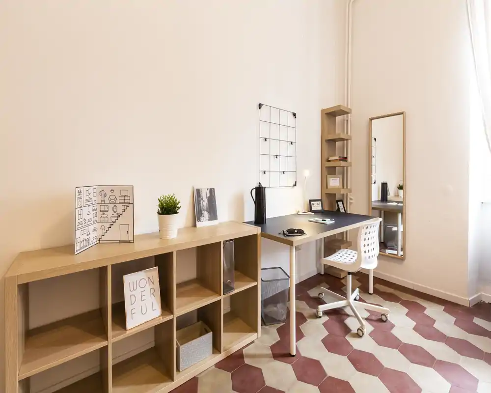 Bright, minimalist living/working corner with a wooden shelving unit, desk and chair beside a large window. Clean lines, warm tones and decorative accents create a calm, functional space ideal for remote work or relaxation.