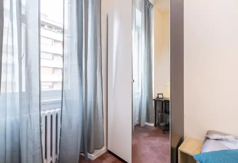 Coin de chambre compact baigné d'une lumière naturelle douce provenant d'une grande fenêtre, rideaux légers, une armoire étroite à porte miroir et un lit partiellement visible au linge simple.