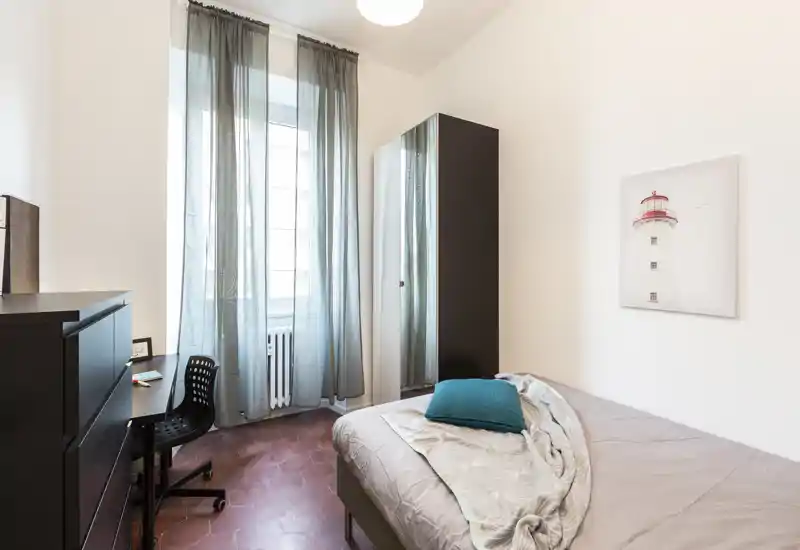 Chambre simple lumineuse et moderne avec lit bien fait, coussin turquoise et voilages légers sur une grande fenêtre ; comprend une armoire et un petit bureau, idéale pour étudiant ou professionnel.