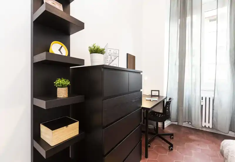 Rincón de dormitorio luminoso y moderno con cómoda oscura, estantes abiertos y un pequeño escritorio junto a la ventana — estilo ordenado con plantas y luz natural.