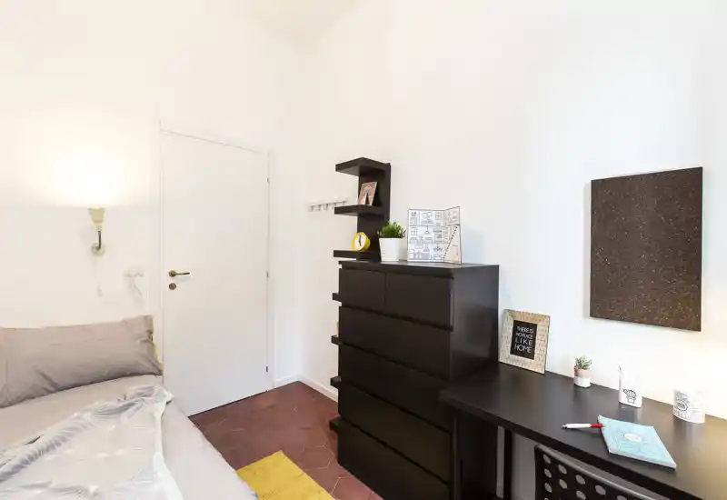 Chambre compacte et lumineuse avec un lit bien fait à gauche, une grande commode noire et un petit bureau décoré — moderne, fonctionnelle et bien éclairée.