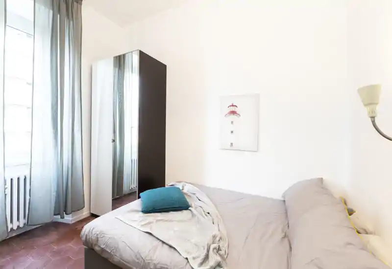 Chambre lumineuse et minimaliste avec un lit double aux tons neutres, un coussin turquoise, une grande armoire miroir et des voilages laissant entrer la lumière naturelle.