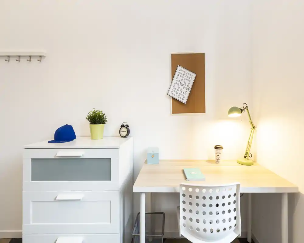 Angolo di lavoro essenziale in un soggiorno luminoso: scrivania in legno chiaro, lampada da scrivania, piccolo cassettone e decorazioni semplici — perfetto per studio o smart working.