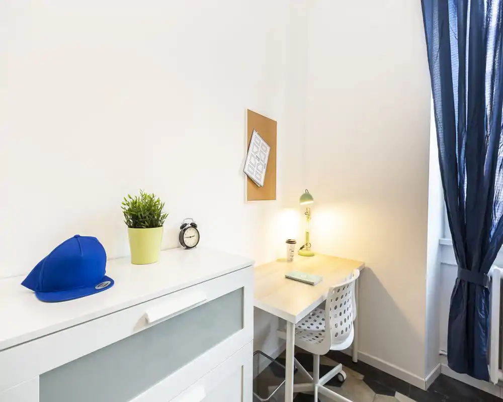 Angolo studio luminoso e minimal con scrivania bianca, sedia traforata, lampada da tavolo e piccolo mobile — spazio pulito e funzionale ideale per studiare o lavorare da remoto.