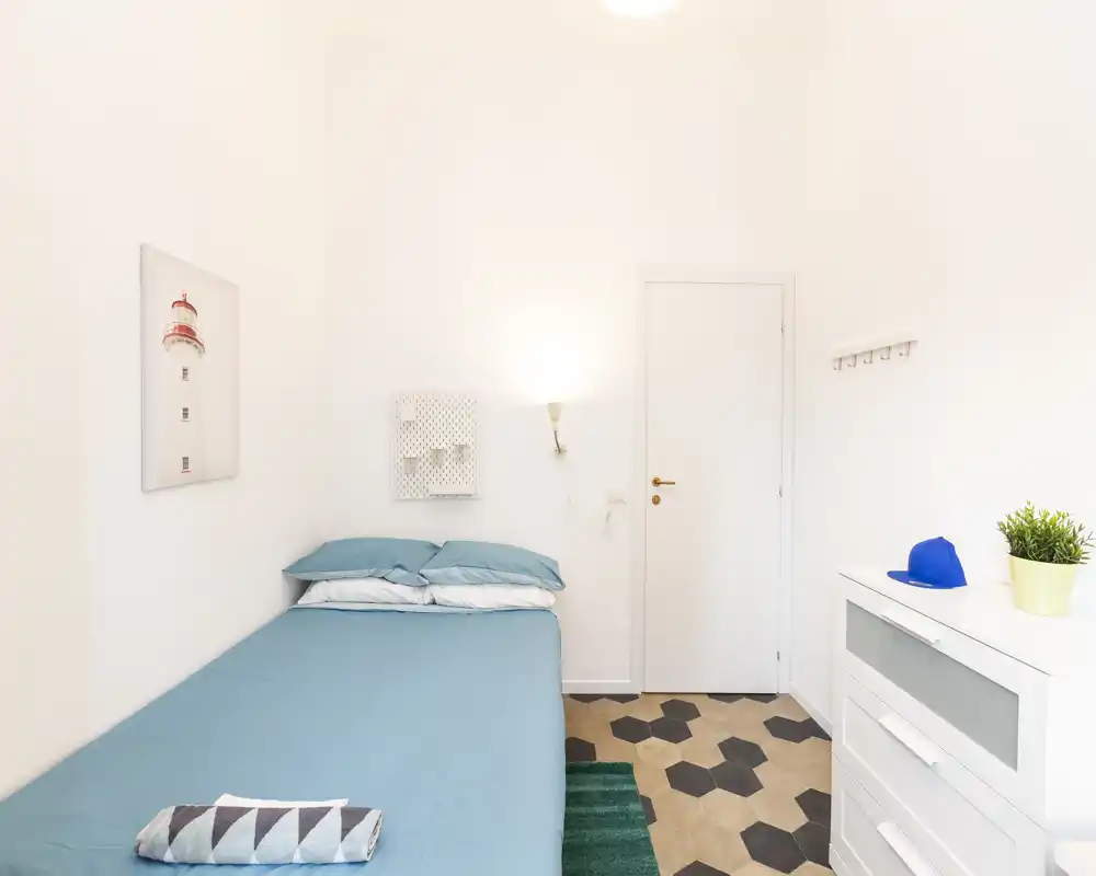 Camera singola luminosa e minimal con letto in biancheria turchese, pareti bianche e pavimento a piastrelle esagonali. Cassettiera compatta, appendini a muro e luce soffusa rendono lo spazio ordinato e accogliente, ideale per brevi soggiorni.