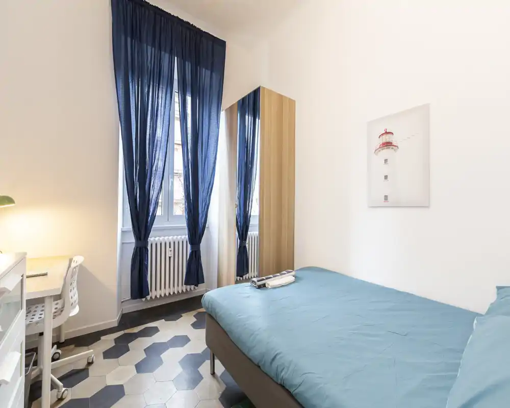 Camera da letto luminosa e compatta con letto matrimoniale con copriletto turchese, armadio alto con specchio, scrivania e ampia finestra con tende blu scuro che lasciano entrare luce naturale.