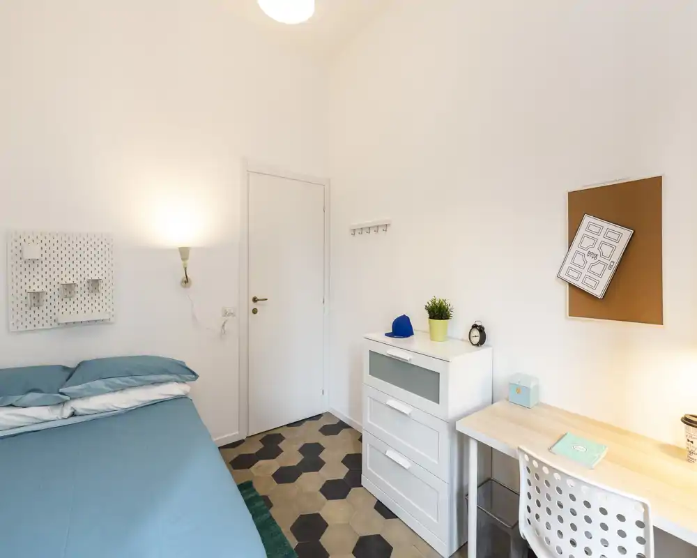 Camera singola luminosa e moderna con copriletto turchese, comò e scrivania compatta — spazio pulito e ben illuminato, ideale per studenti o soggiorni brevi.