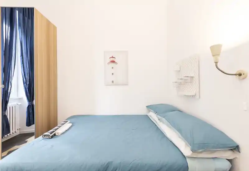 Chambre lumineuse et minimaliste avec un lit double en linge bleu-vert, une lampe murale et une décoration murale discrète. La lumière naturelle passe à travers des rideaux bleu marine, apportant une atmosphère calme et aérée, idéale pour les locations.