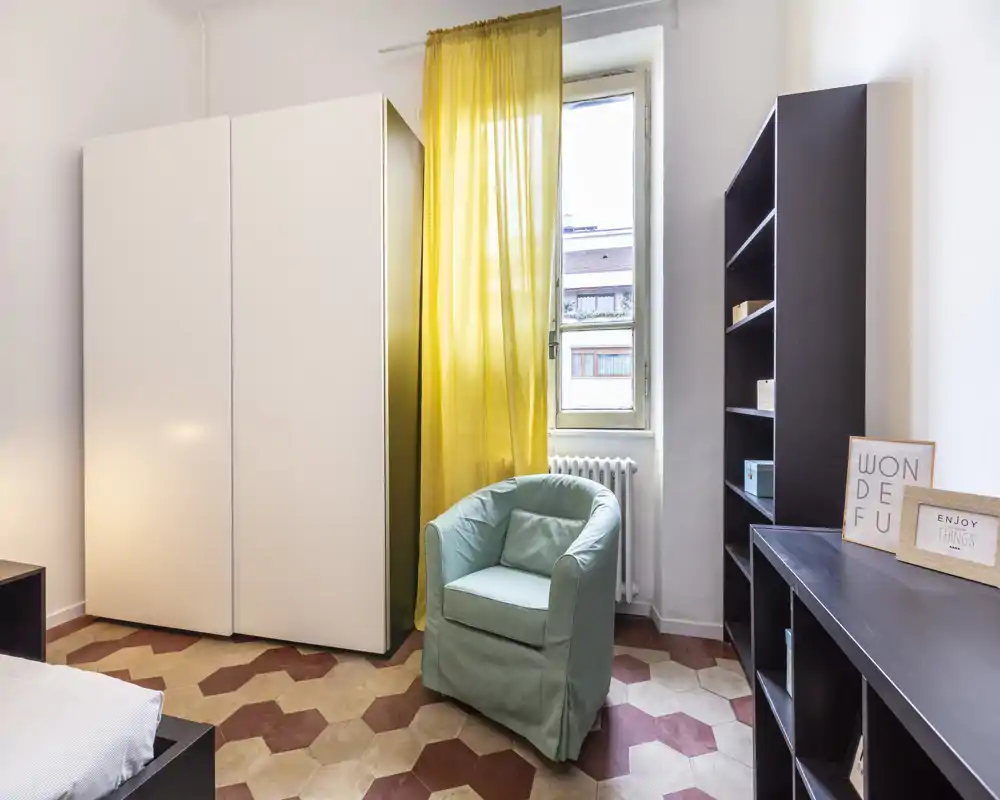 Rincón de dormitorio luminoso con armario, butaca y estantería. Una cortina amarilla y el suelo con dibujo aportan carácter mientras la luz natural entra por la ventana.