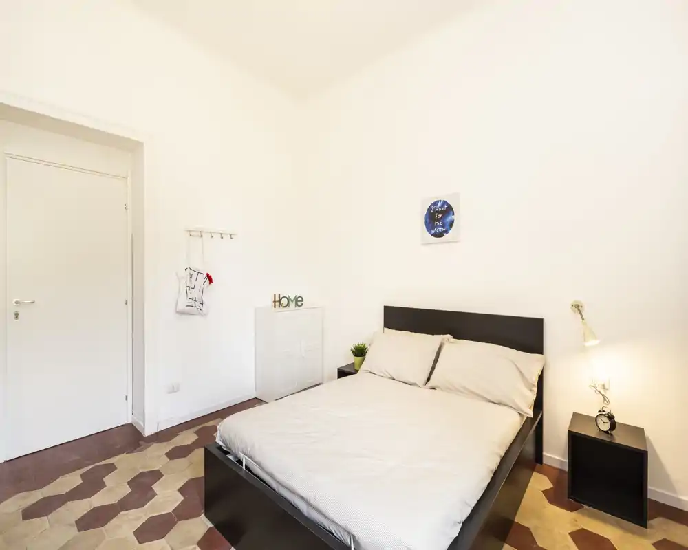 Dormitorio doble luminoso y minimalista con estructura de cama oscura, mesita con lámpara y suelo de baldosas decorativas — limpio y acogedor para alquileres temporales.