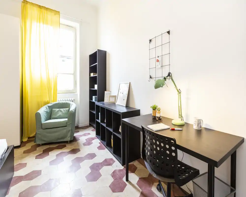 Habitación luminosa y multifuncional con escritorio, estanterías y un sillón cómodo junto a la ventana con cortinas amarillas; ideal para oficina en casa o habitación de estudiante.