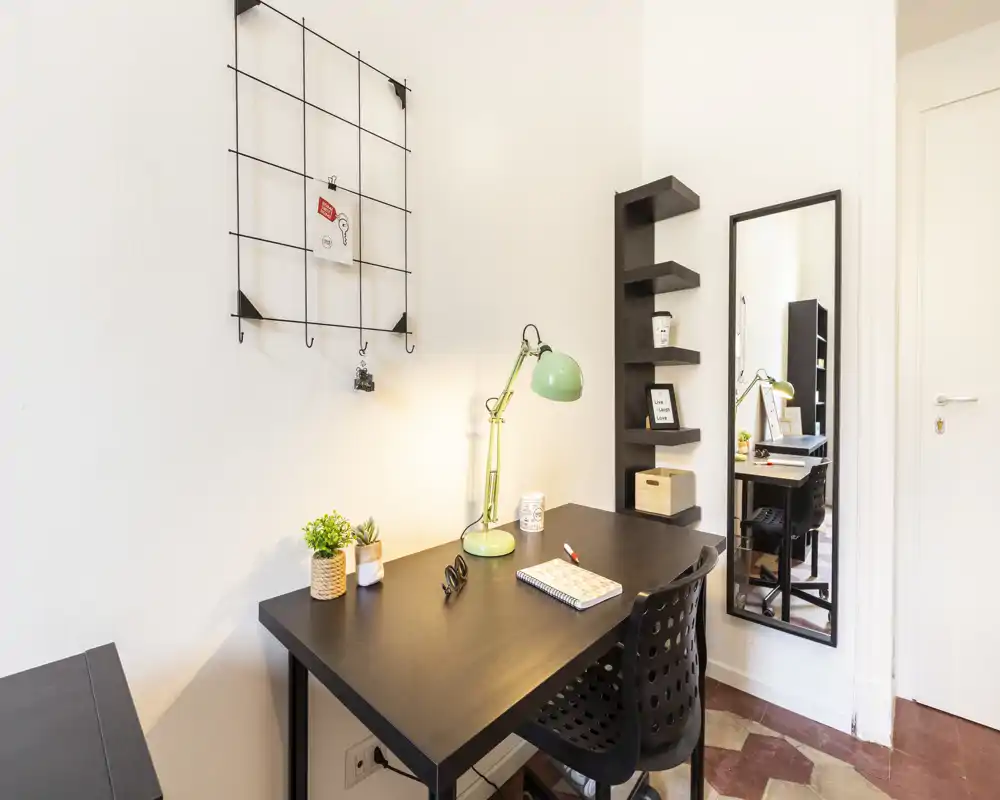 Rincón de estudio moderno y luminoso con escritorio negro, lámpara verde, silla y estantería decorativa — un espacio de trabajo compacto y bien iluminado, ideal para teletrabajo.