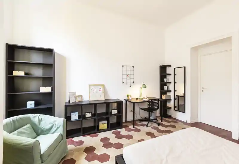 Dormitorio luminoso y moderno con cama parcialmente visible, escritorio con lámpara, estanterías y un sillón cómodo. Suelo de baldosas con patrón y paredes neutras, ideal para anuncios que destaquen el espacio de trabajo y la sencillez acogedora.