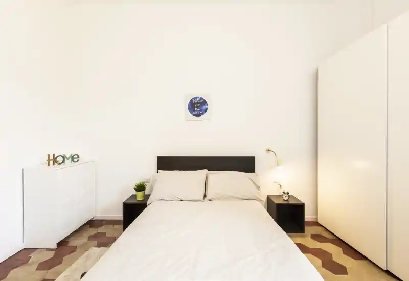 Dormitorio luminoso y minimalista con cama doble centrada, mesitas, lámpara y armario: líneas limpias y suelos con patrón cálido que crean un ambiente acogedor.
