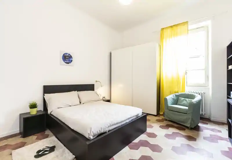 Dormitorio luminoso y ordenado con cama doble, armario y un cómodo sillón junto a la ventana soleada.