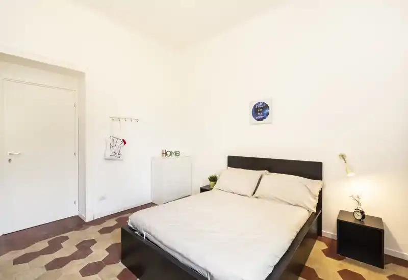 Dormitorio doble luminoso y minimalista con estructura de cama oscura, mesita con lámpara y suelo de baldosas decorativas — limpio y acogedor para alquileres temporales.