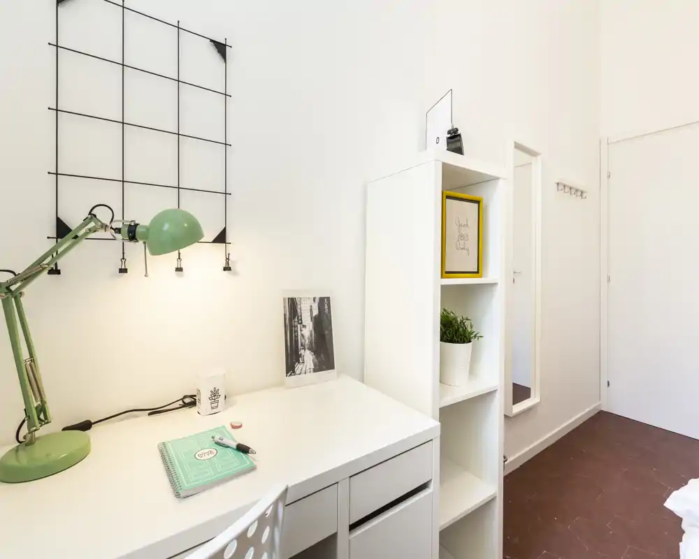 Rincón de dormitorio bien iluminado con escritorio blanco, lámpara verde y estantería; decoración moderna y minimalista con un vislumbre de la cama en el borde derecho.