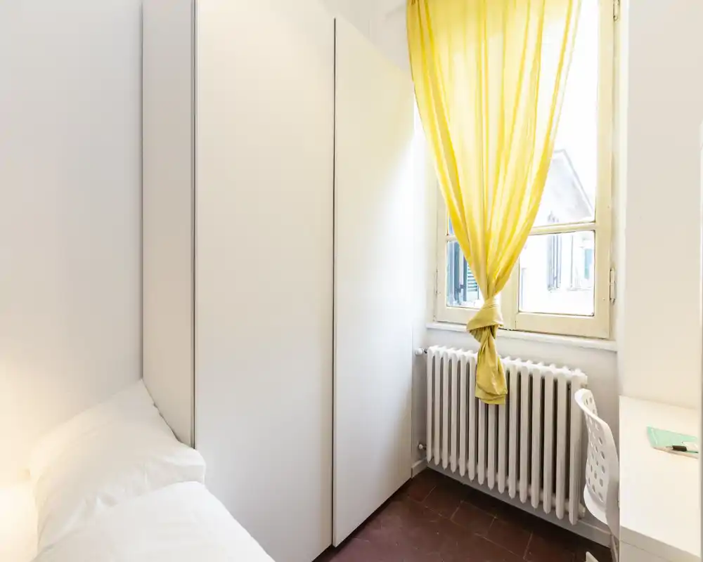 Rincón de dormitorio compacto con cama blanca parcialmente visible, armario alto, escritorio y una ventana luminosa con una cortina amarilla que añade un toque alegre.