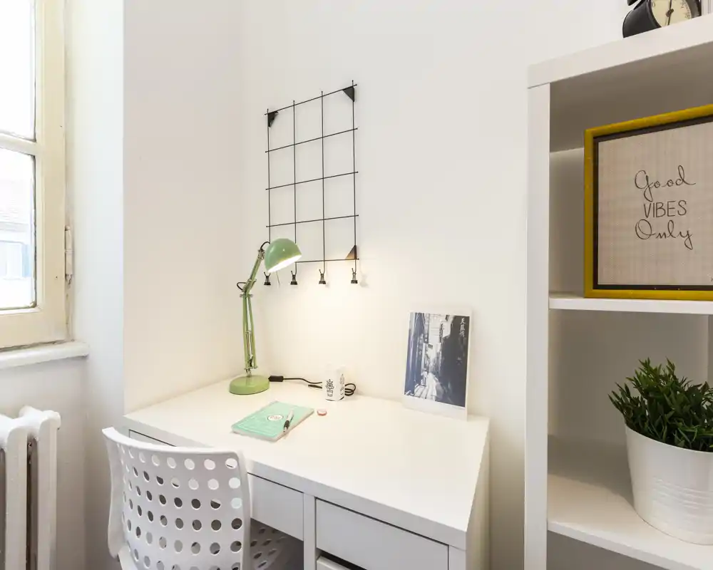 Rincón de estudio luminoso y ordenado en el dormitorio con escritorio blanco, lámpara verde y estanterías—ideal como zona de trabajo.