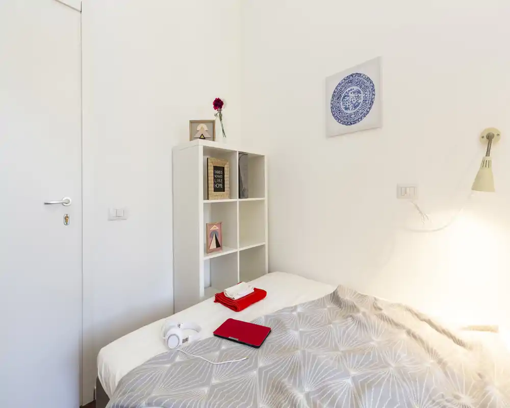Dormitorio individual luminoso y minimalista con cama bien hecha, edredón de colores claros, estantería junto a la cama y decoración sencilla en la pared — bien iluminado y ordenado para el anuncio.