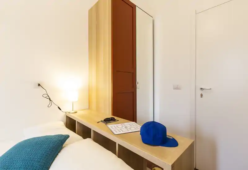 Dormitorio moderno y luminoso que muestra el borde de la cama, una lámpara de noche y un armario con espejo — acogedor y bien iluminado, ideal para anuncios.