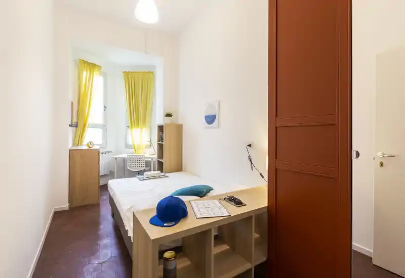 Dormitorio moderno y luminoso con cama doble bien hecha, un pequeño escritorio junto a la ventana y cortinas amarillas que dejan pasar la luz natural.