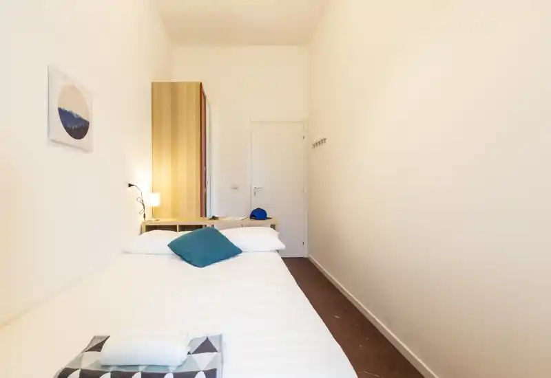 Habitación compacta con cama individual bien hecha, lámpara de mesita, armario y tonos neutros que crean un espacio luminoso, minimalista y acogedor.
