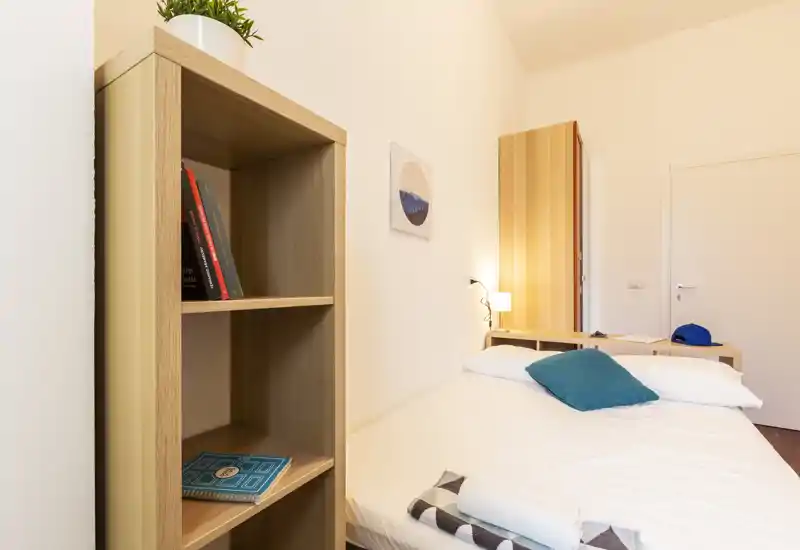 Dormitorio bien iluminado y ordenado con cama doble, lámpara de mesita y muebles de madera clara que crean un ambiente cálido y minimalista.