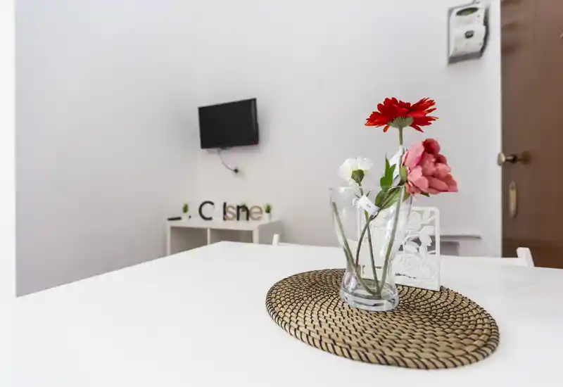 Zona pranzo minimal luminosa con tavolo bianco, sottopiatto intrecciato e centrotavola floreale; sullo sfondo una TV a parete e un piccolo mobiletto.