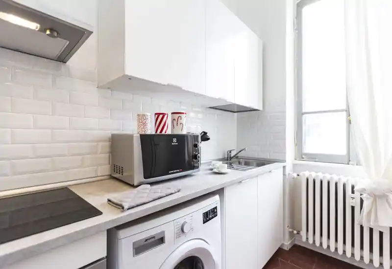 Cucinotto moderno e luminoso con mobili bianchi, piano cottura a induzione, microonde, lavello e lavatrice sotto il piano — perfetto per appartamenti in città.
