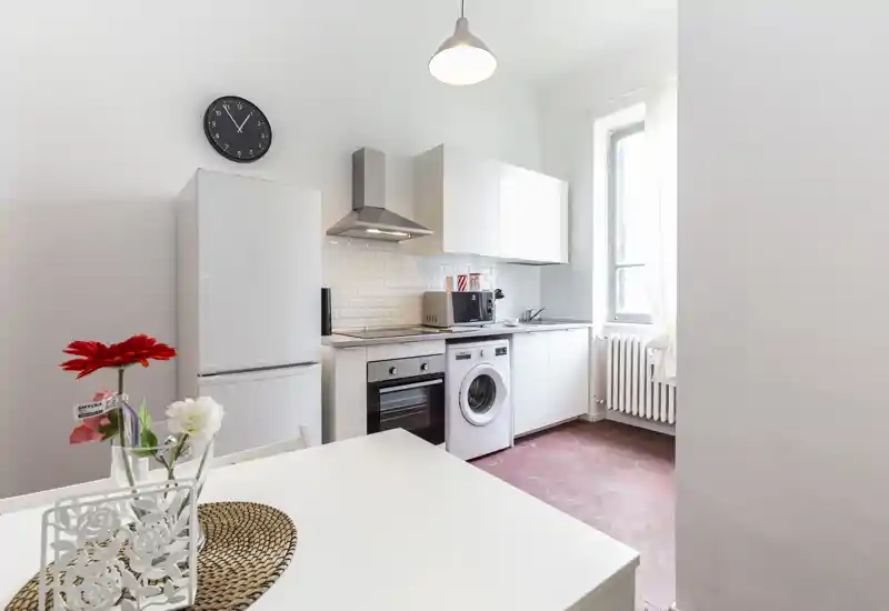 Cucina compatta e luminosa con pensili bianchi, forno, frigorifero e lavatrice integrata sotto il piano. Un piccolo tavolo da pranzo con una decorazione semplice e una finestra che illumina lo spazio rendono l’ambiente ordinato e funzionale.