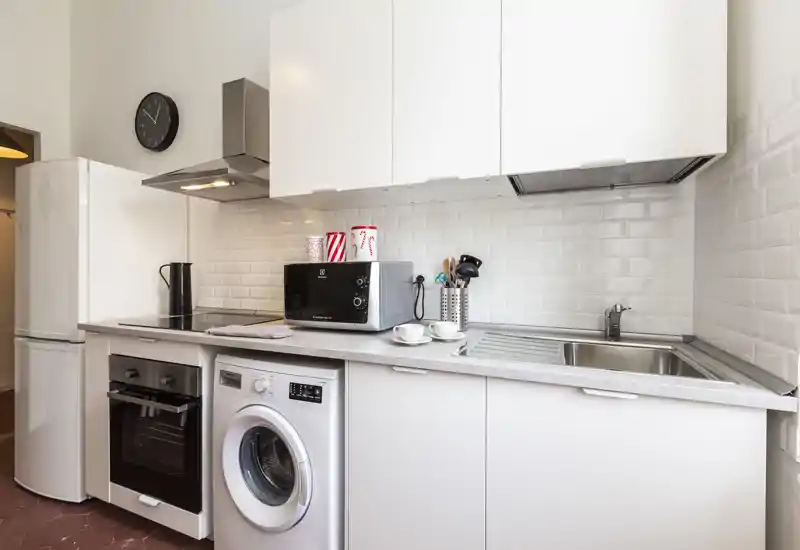 Cucina moderna compatta con mobili bianchi, forno incorporato, lavatrice, microonde e lavello in acciaio; piastrelle a metrò pulite e arredo essenziale ben illuminato, ideale per affitti brevi.