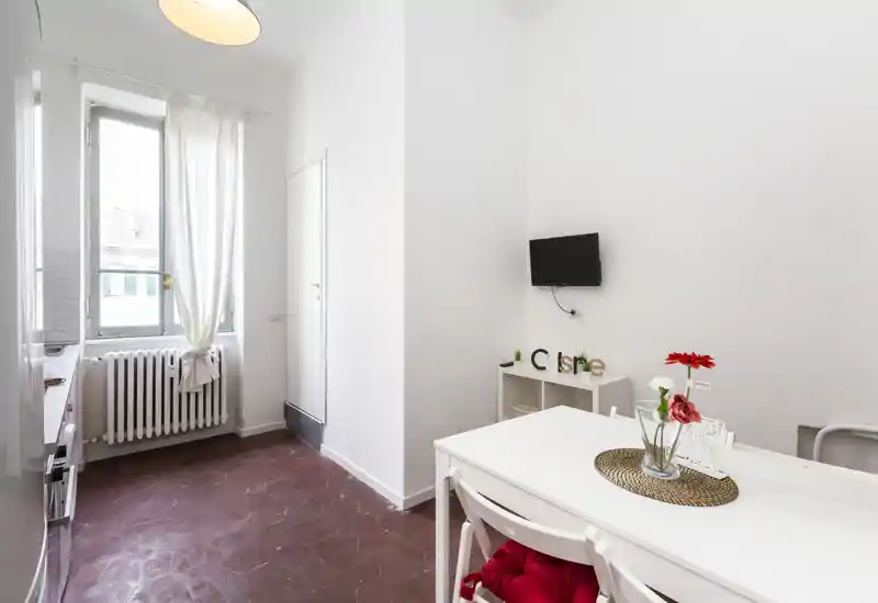 Area giorno luminosa e minimale con tavolo bianco, sedie e una piccola TV a parete; luce naturale da una finestra alta e decorazioni semplici creano un ambiente pulito e accogliente.