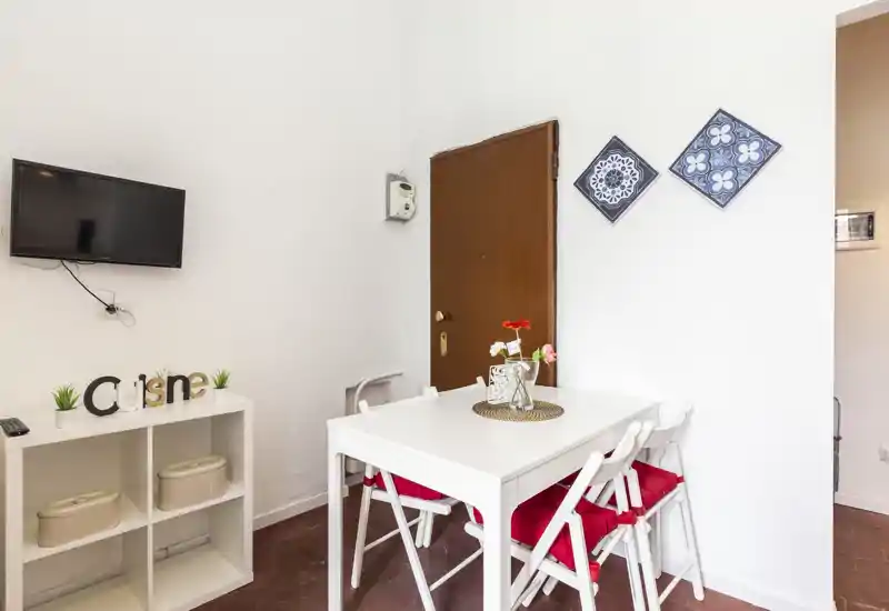 Angolo pranzo luminoso e ordinato con tavolo bianco, quattro sedie con cuscini rossi e piccolo centrotavola floreale — ideale per pasti informali in un appartamento compatto.