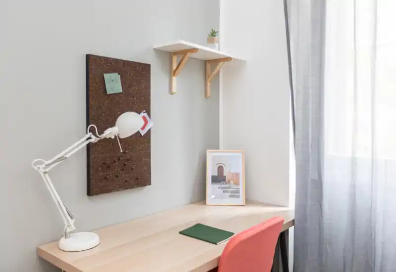 Neat workspace corner with a light wooden desk, adjustable white desk lamp, cork notice board and a coral chair beside a bright window with sheer curtains.