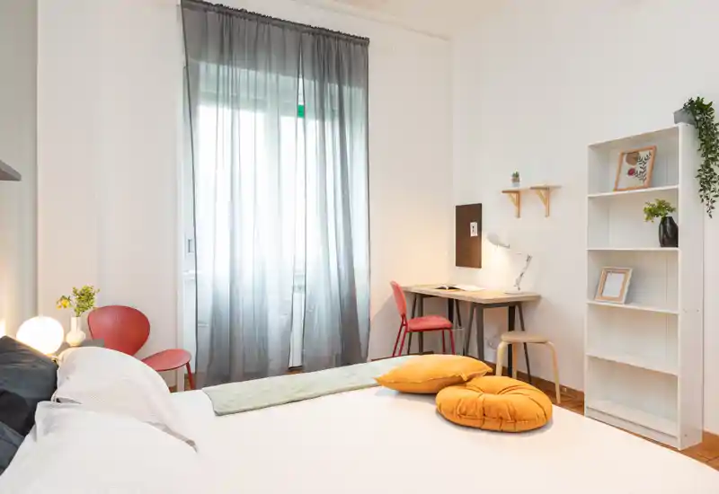 Camera da letto luminosa e moderna con un grande letto in primo piano, cuscini color senape e una piccola scrivania vicino alla finestra: perfetta per soggiorni confortevoli o per lavorare da casa.
