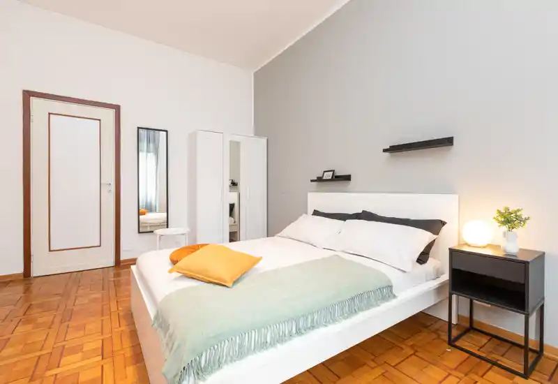 Camera da letto luminosa e moderna con letto matrimoniale, comodino e armadio. Arredi bianchi e tessili morbidi creano un ambiente fresco e accogliente, perfetto per annunci.