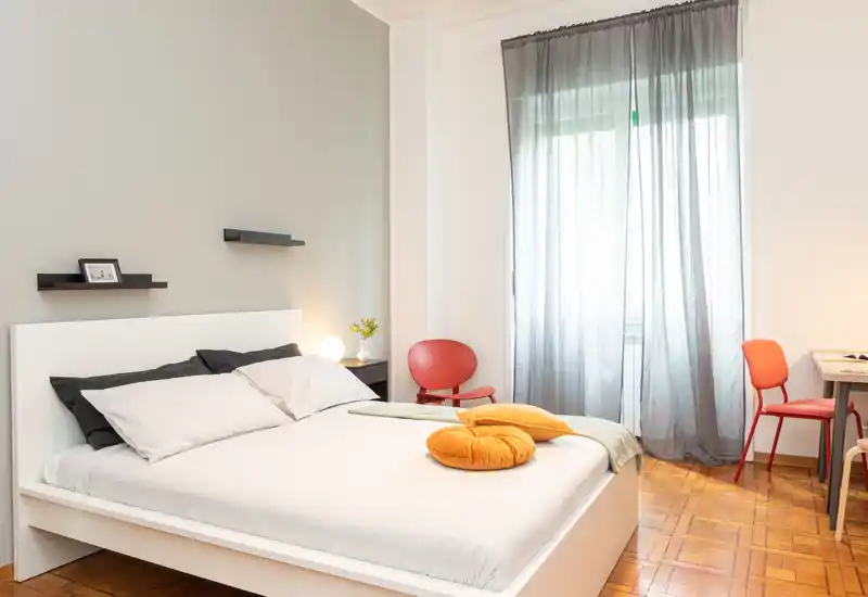 Camera da letto moderna e luminosa con letto bianco, biancheria morbida e cuscini decorativi, luce naturale abbondante da una grande finestra e arredo minimalista.