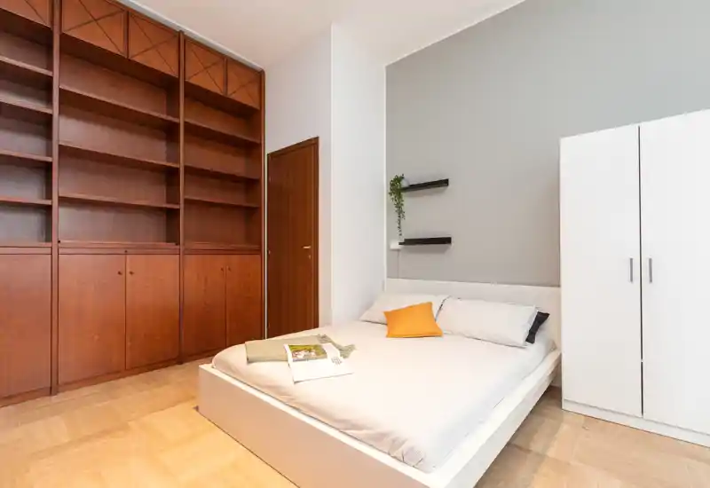 Dormitorio amplio y luminoso con cama baja, estanterías empotradas de madera y armario blanco; tonos neutros y un cojín de acento que crean un ambiente acogedor.