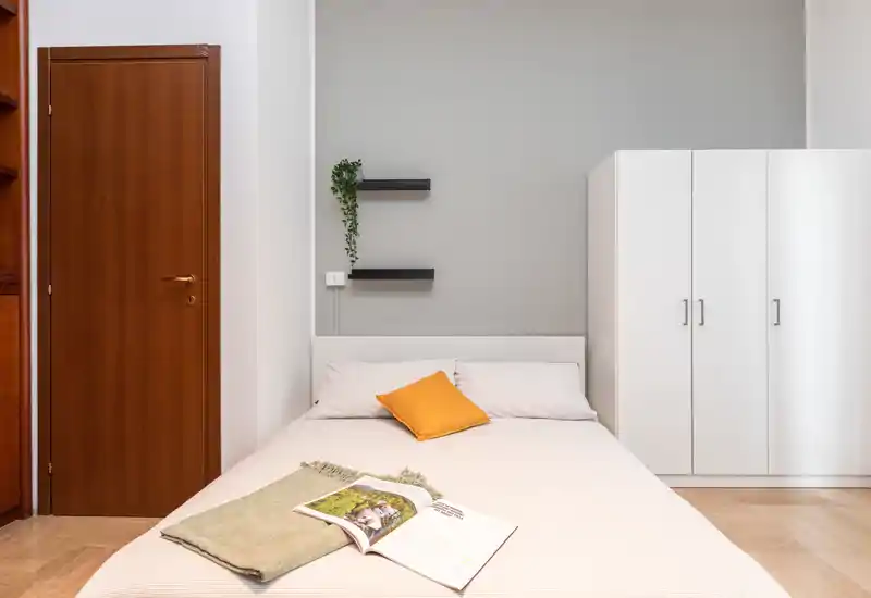 Dormitorio luminoso y minimalista con cama doble centrada, cojín mostaza y armario a la derecha; líneas limpias y tonos neutros que crean un ambiente tranquilo.