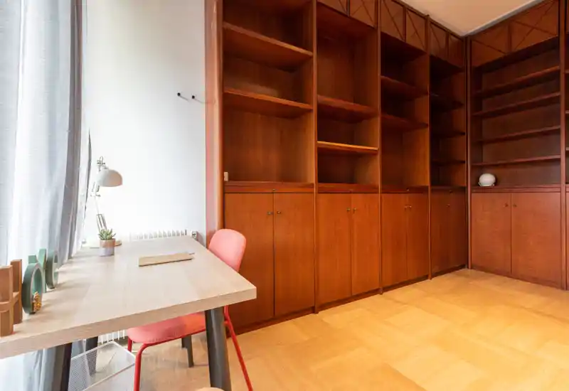 A bright study/corner living area with a simple desk and chair beside tall built-in wooden shelving units. Natural light fills the space and warm wood tones create a calm, functional atmosphere.