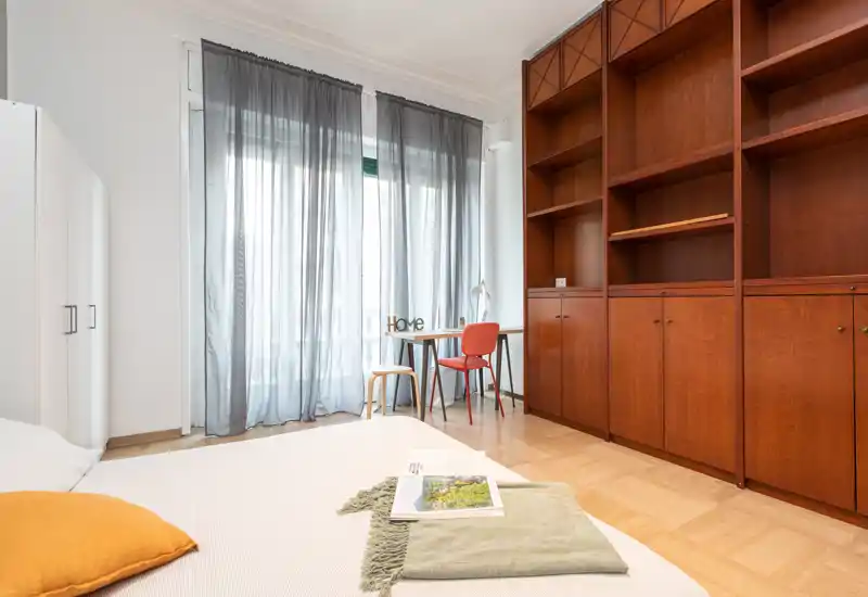 Dormitorio luminoso y bien compuesto con cama baja en primer plano, amplia estantería de madera y escritorio junto a una ventana alta con cortinas translúcidas que filtran la luz natural.