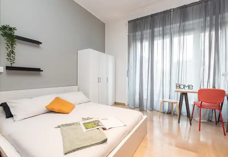 Dormitorio moderno y luminoso con una cama grande, ropa de cama suave y luz natural de una ventana hasta el suelo; armario sencillo y un pequeño escritorio que crean un espacio funcional y acogedor.