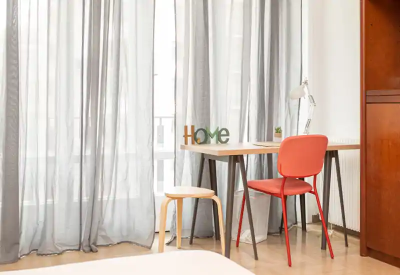 Bright bedroom corner with a small desk and red chair by sheer curtains; minimal glimpse of the bed in the foreground suggests this is a sleeping area with a practical workspace.